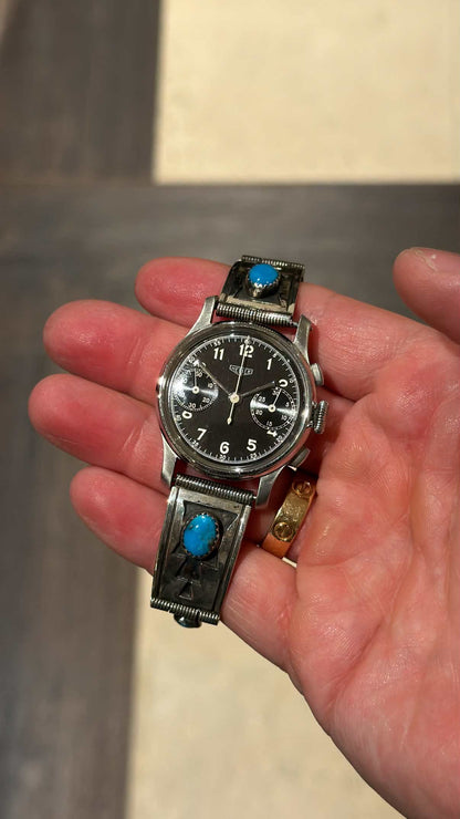 Front view of 1940s Heuer black dial chronograph on stamped sterling silver turquoise bracelet held in palm