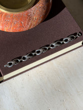 Twisted link bracelet laid flat along a brown book beneath a red stone ashtray, the patinated figure eight links in a row