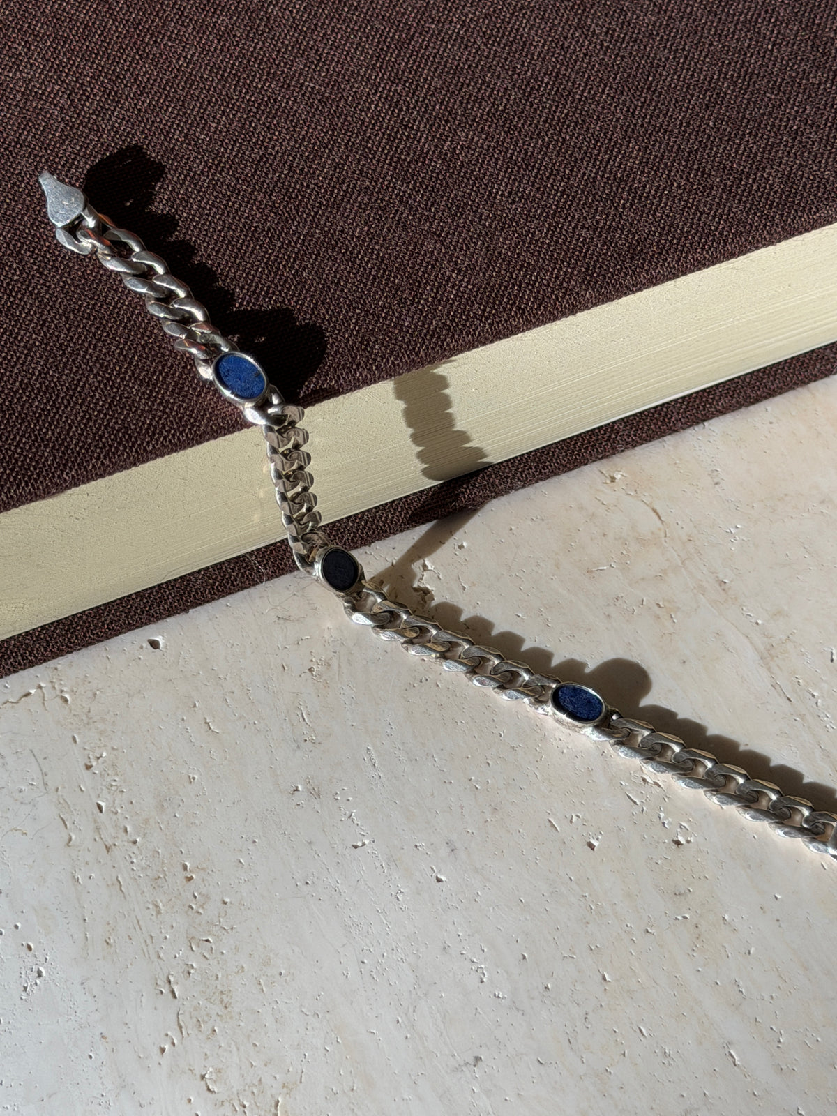 Lapis lazuli curb bracelet draped from a brown book onto travertine in raking sunlight, its box clasp at one end