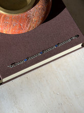 Overhead view of the curb bracelet with three blue lapis ovals laid straight on a brown book below a red stone ashtray with brass well