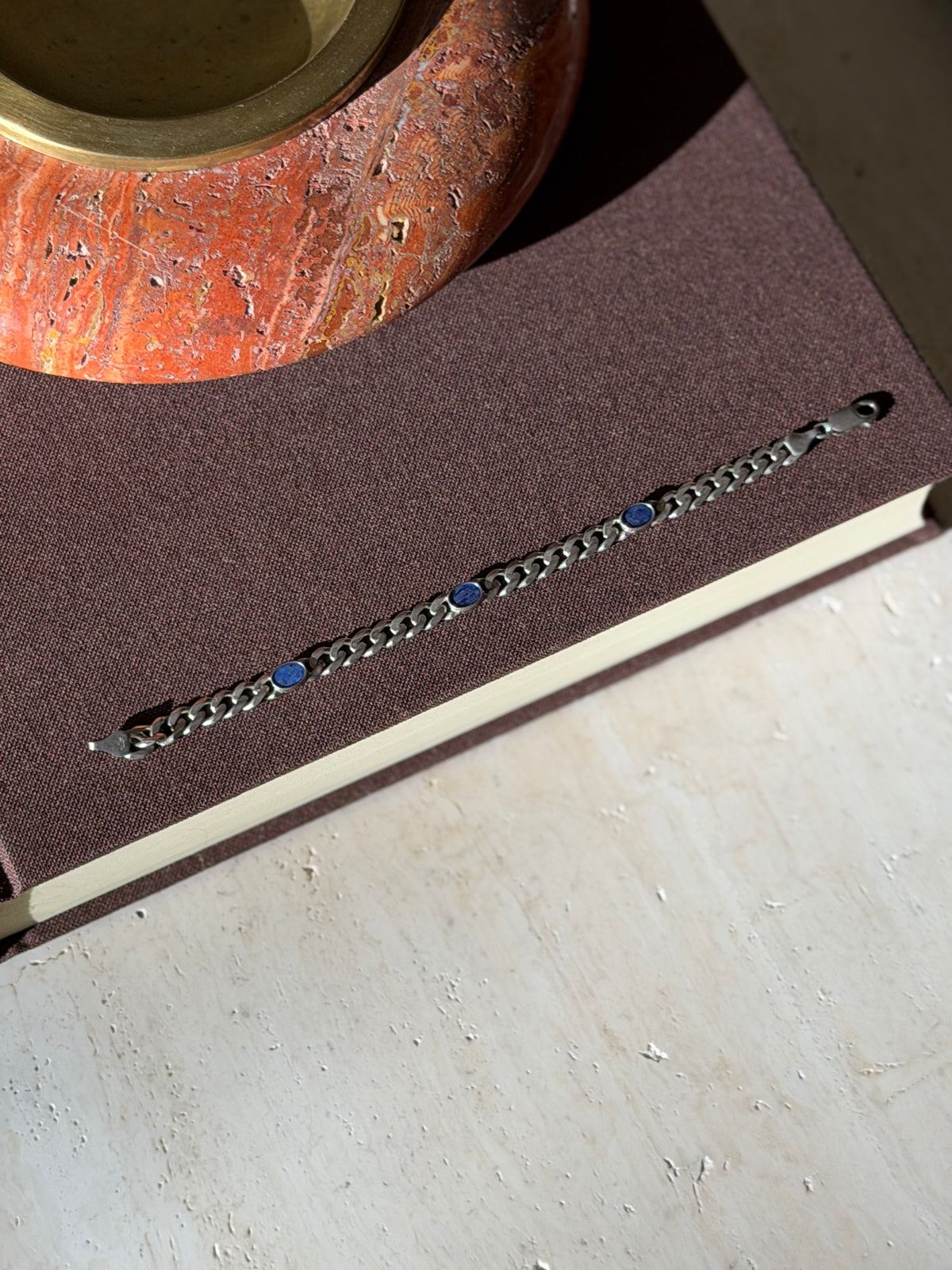 Overhead view of the curb bracelet with three blue lapis ovals laid straight on a brown book below a red stone ashtray with brass well