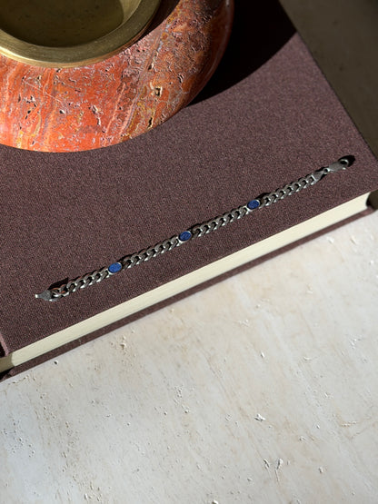 Overhead view of the curb bracelet with three blue lapis ovals laid straight on a brown book below a red stone ashtray with brass well