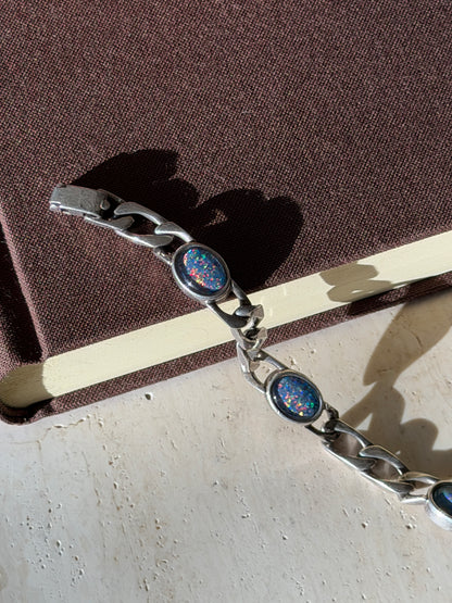 Close view of oval opal cabochons glinting with red, green and blue fire, the bracelet and box clasp draped over a brown book edge