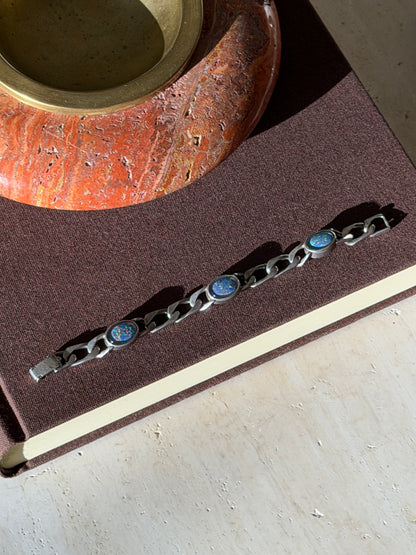 Overhead view of the curb link bracelet with three oval opals on a brown book, a red stone ashtray above in afternoon light