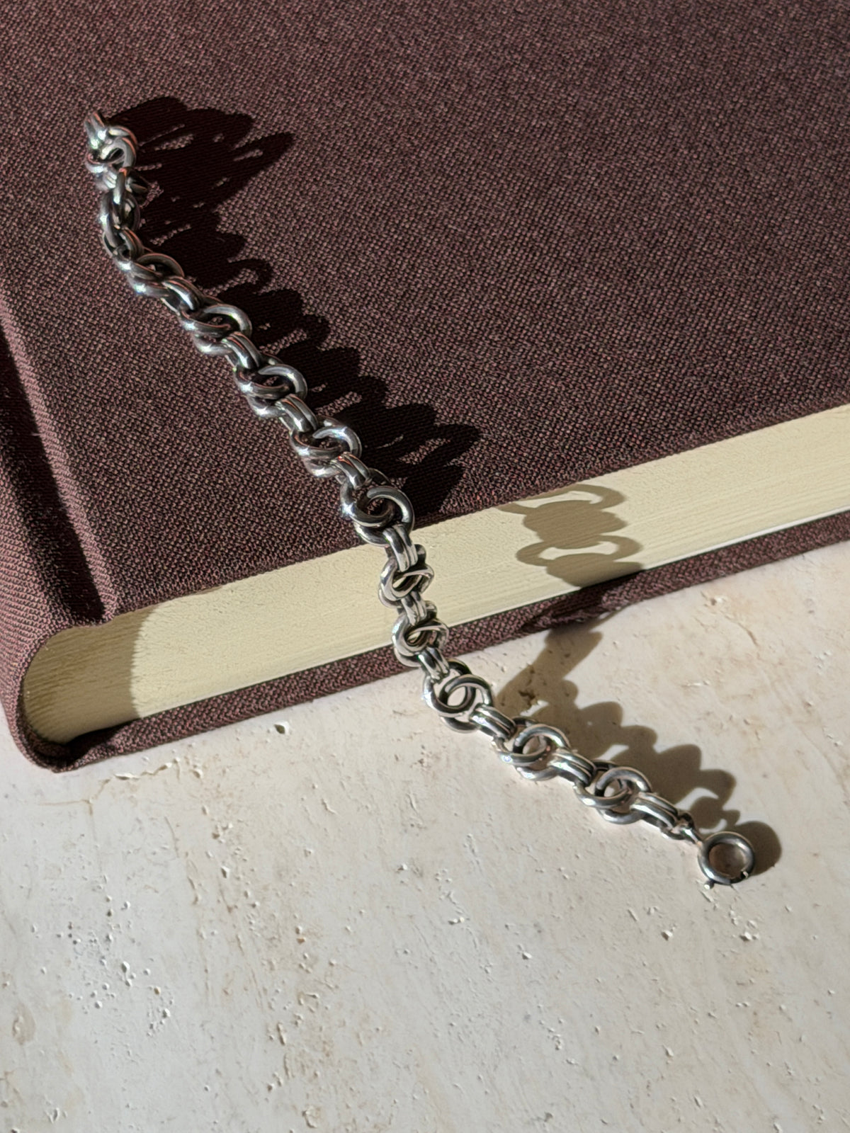 Ring link silver bracelet draped from a brown book onto travertine, its spring ring clasp and looping shadows in sunlight