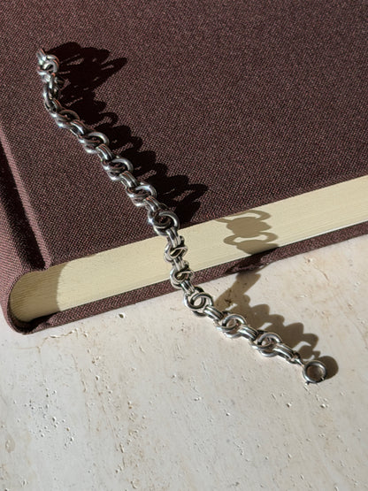 Ring link silver bracelet draped from a brown book onto travertine, its spring ring clasp and looping shadows in sunlight