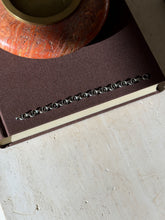 Overhead view of the double ring and bar link bracelet laid along a brown book below a red stone ashtray