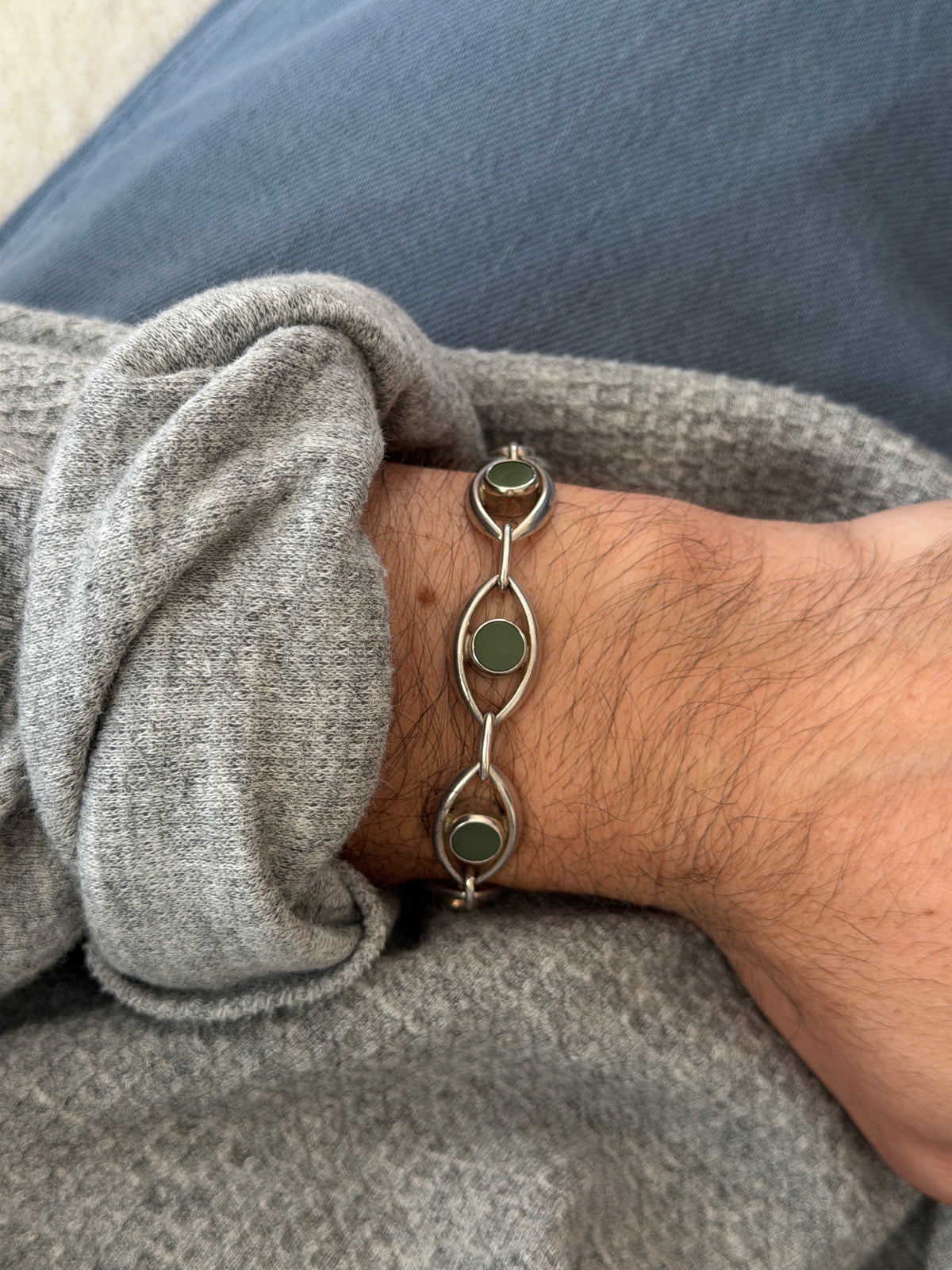 Vintage German sterling silver bracelet of open almond shaped eye links, each centered with a round green jade disc, worn on the wrist over a gray sweater and blue denim