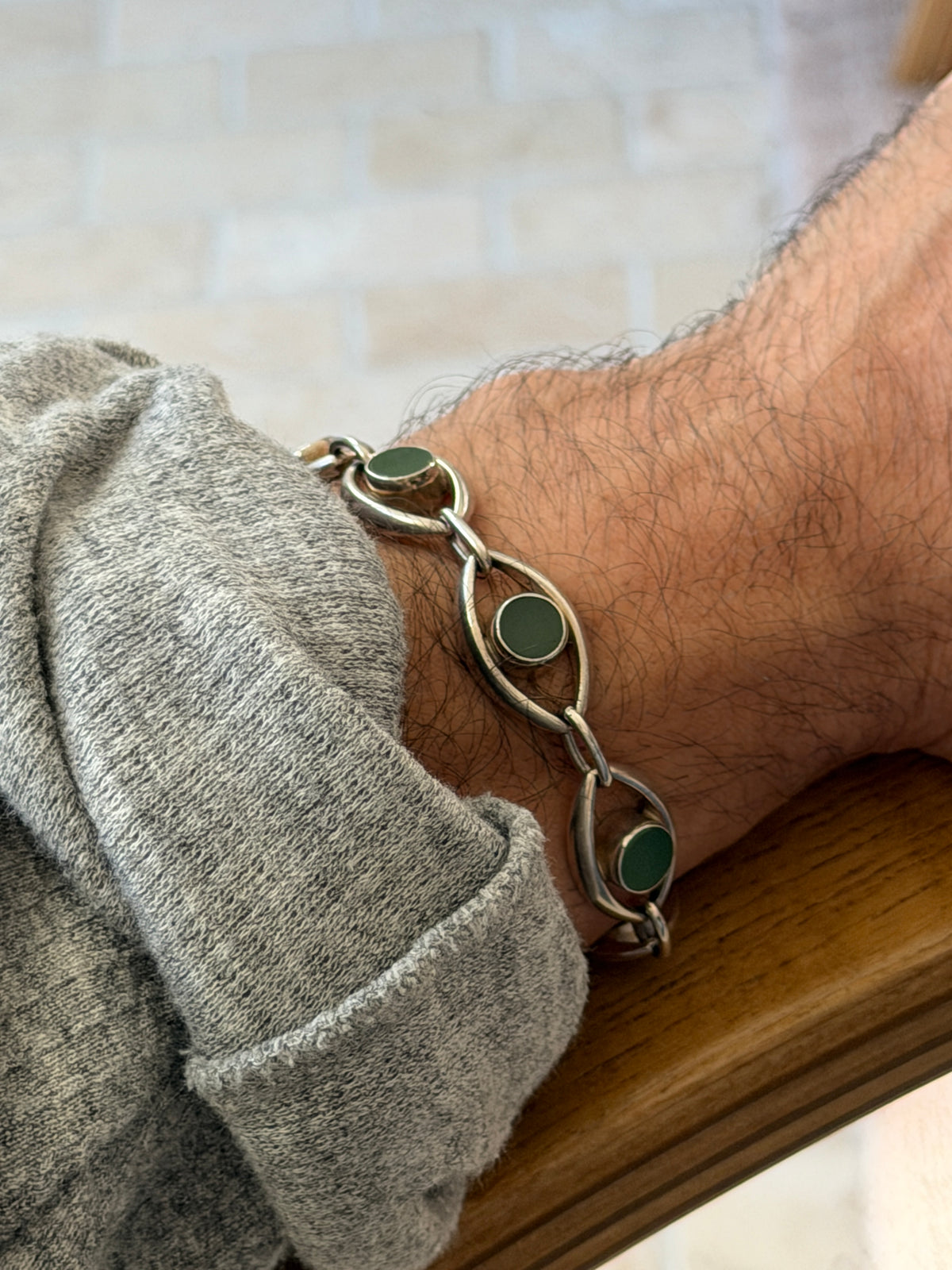 Jade and silver eye link bracelet on the wrist resting on a wooden chair arm, deep green stones against a soft brick wall