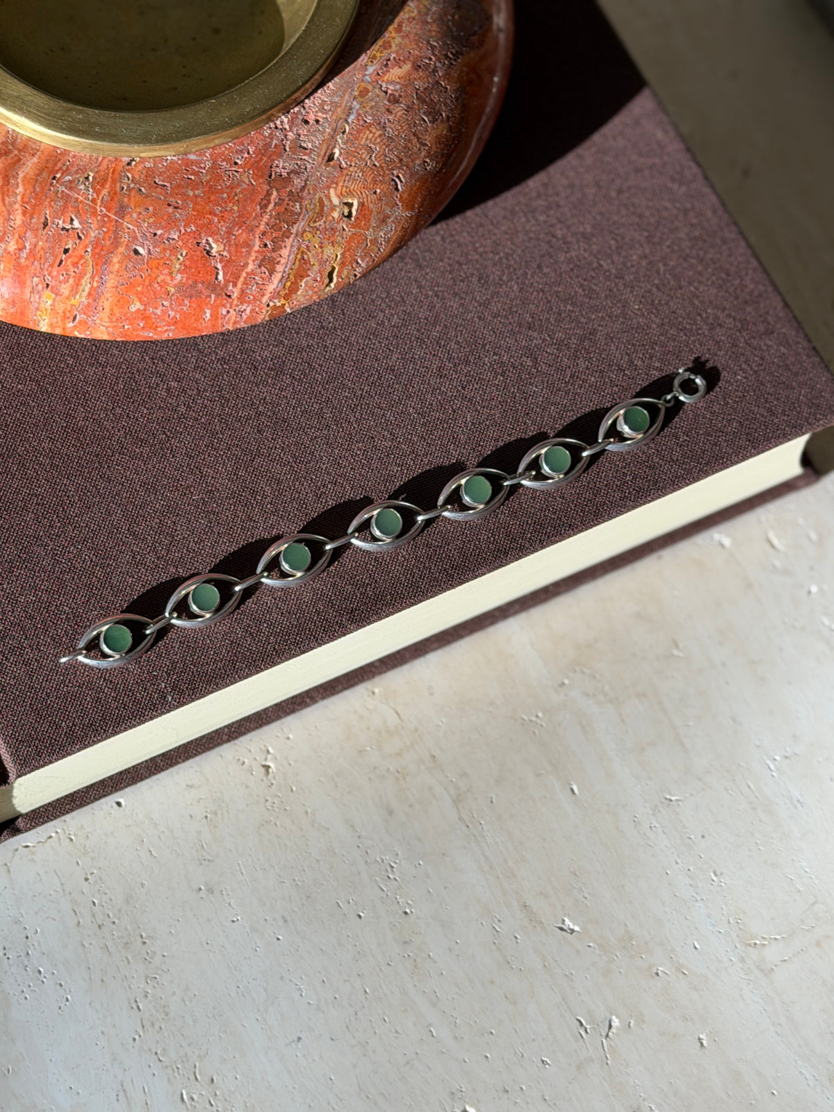 Overhead view of the sterling bracelet with seven jade eye links laid straight on a brown book beneath a red stone ashtray with brass well