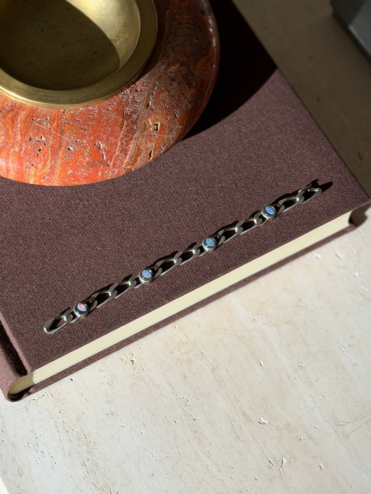 Overhead view of the opal link bracelet laid along a brown book beneath a red stone ashtray, four round opals spaced along the chain