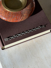 Overhead view of the sterling ring and bar bracelet laid straight on a brown book beneath a red stone ashtray with brass well