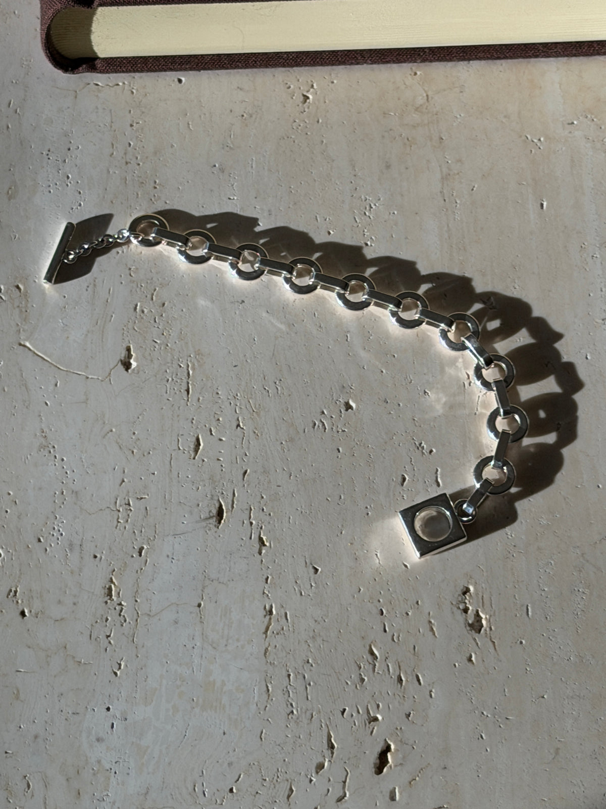 Sterling round link bracelet curved across travertine in raking sunlight, a T bar on a short chain at one end and a square toggle ring at the other
