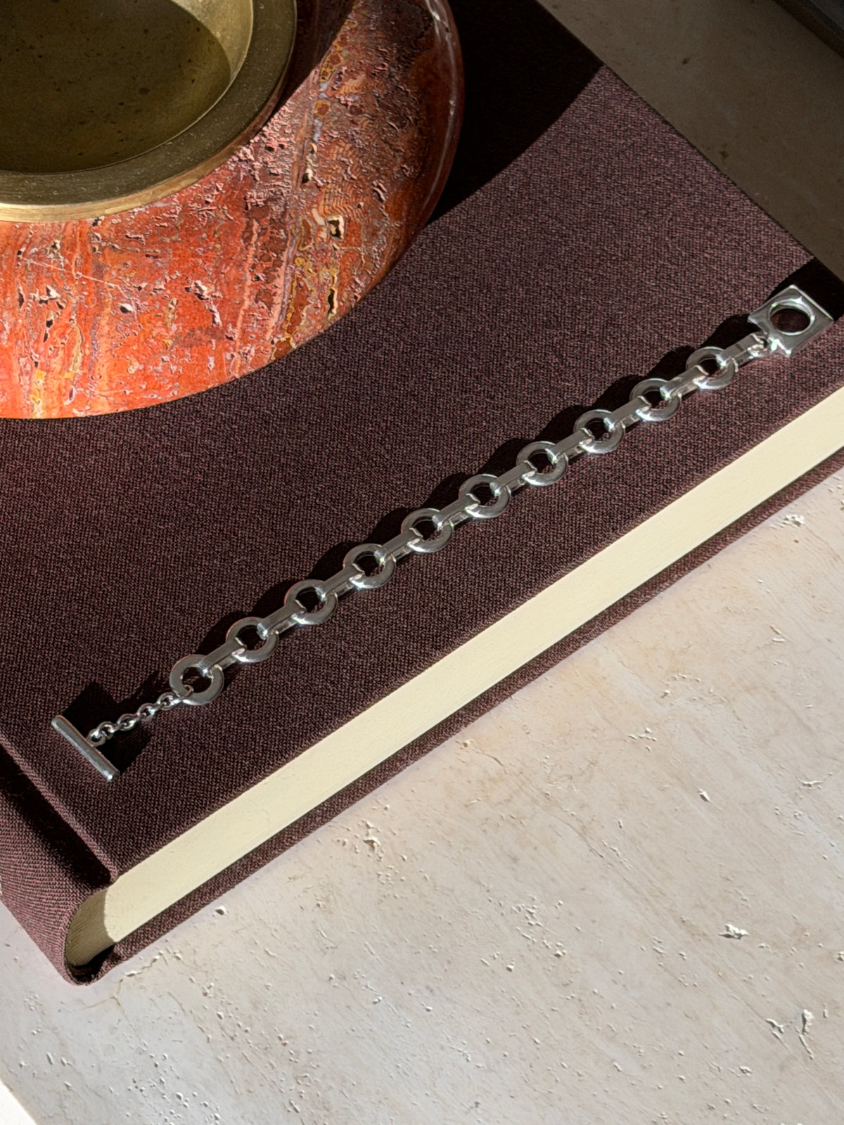 Overhead view of the round link and square toggle bracelet laid straight on a brown book beneath a red stone ashtray with brass well