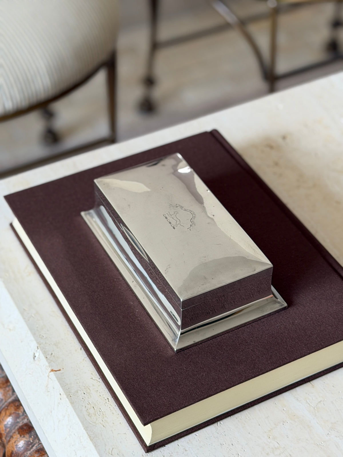 Sterling silver box on a brown book atop travertine, a striped chair behind, the crest catching light on the polished lid