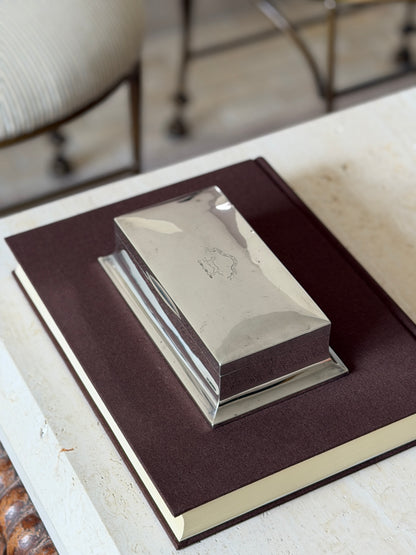 Sterling silver box on a brown book atop travertine, a striped chair behind, the crest catching light on the polished lid