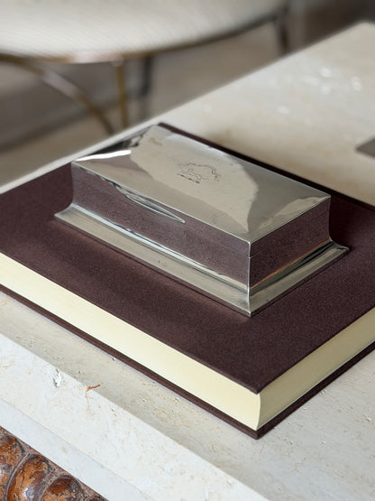 Angled view of the Walker and Hall box on a brown book, the hinged lid and splayed rectangular base in daylight