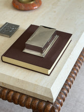 High angle view of the silver box on a brown book at the travertine table edge, a red stone ashtray and pewter box beyond